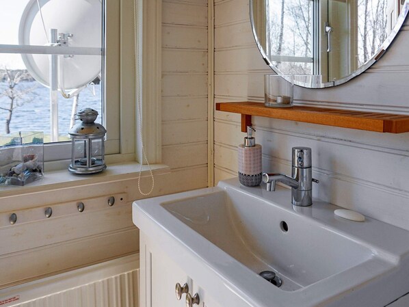 A bathroom with a large sink, a mirror, a window with a view of the sea, and a shelf with a bottle and a lantern.
