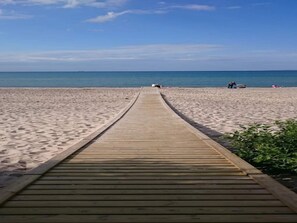 Une promenade en bois mène à une plage de sable fin, sous un ciel bleu et dégagé.