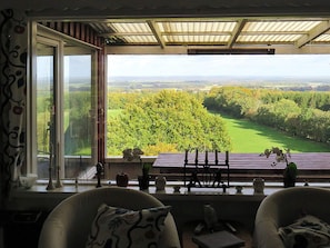 A room with a large window overlooking a green landscape, a wooden ceiling, and two chairs with cushions.