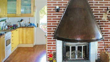 A kitchen with wooden cabinets and a stove, a brick fireplace with a metal hood, and a window with blinds.