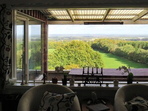 Ein Zimmer mit großem Fenster und Blick auf eine grüne Landschaft, Holzboden und einer Sitzecke mit Kissen.