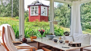 Une véranda avec une table en bois, des chaises et une vue sur une maison rouge et la verdure.