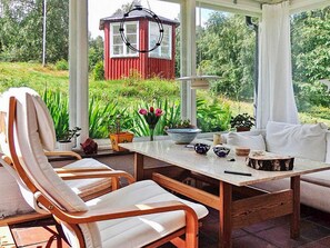 A conservatory with a wooden table, chairs, and a view of a red house and greenery.