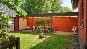 A covered outdoor area with a red wall, a wooden pergola, and a small garden with a tree and a water feature. - 10 Person Holiday Home in Nexo (Nexø)
