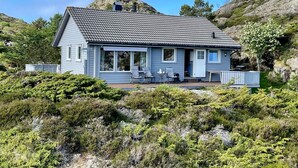 A single-story house with a grey tiled roof, a covered porch with outdoor seating, and a view of a rocky hillside. - 4 Star Holiday Home in Espevaer (Bomlo)