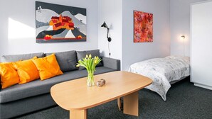Ein modernes Hotelzimmer mit grauem Sofa, einem Holztisch mit Blumenvase, einem Bett mit weiß-grauem Muster sowie zwei Kunstwerken an den Wänden.