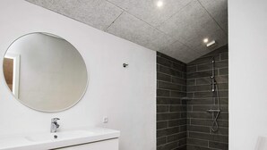 A modern bathroom with a white vanity, a round mirror, a shower with dark tiles, and a white toilet.