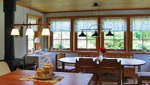 Un coin repas chaleureux avec une table et des chaises en bois, un poêle et de grandes fenêtres ornées de rideaux à motifs floraux.