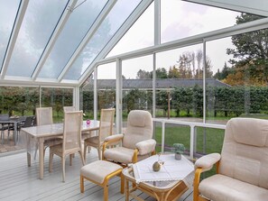 Ein Wintergarten mit Korbmöbeln und einem Tisch, großen Fenstern sowie Blick auf Garten und Haus.