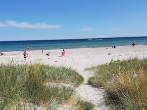 En sandstrand med folk som promenerar och sitter, en klar himmel och havet i bakgrunden.