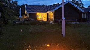 A house with a fire pit in the foreground, a wooden fence, and a well-lit interior. - 4 Star Holiday Home in Karrebaeksminde (Karrebæksminde)