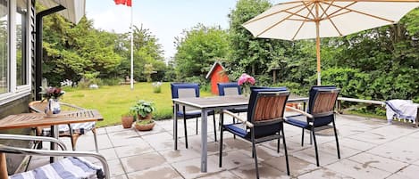 An outdoor patio with a table and chairs, a flag, and a garden in the background.