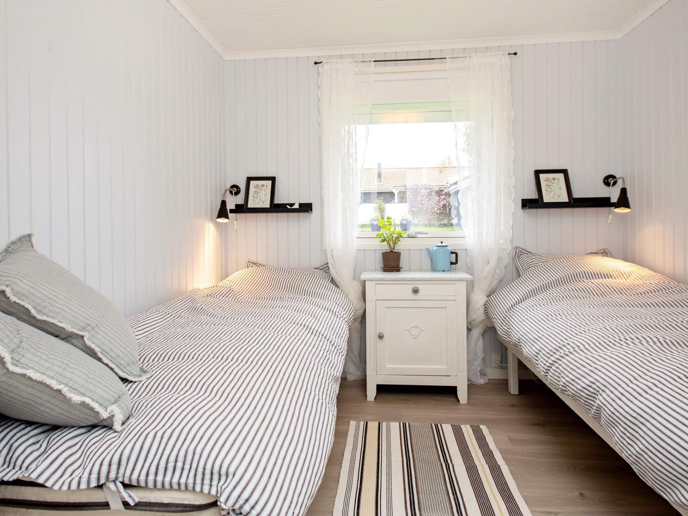 A bedroom with two beds, a nightstand, a window with curtains, and wall-mounted lamps.