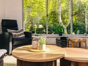A modern living room with a large window, a black leather sofa, a wooden coffee table with decorative items, and a view of a lush garden. - 6 Person Holiday Home in Hadsund (Hadsund)