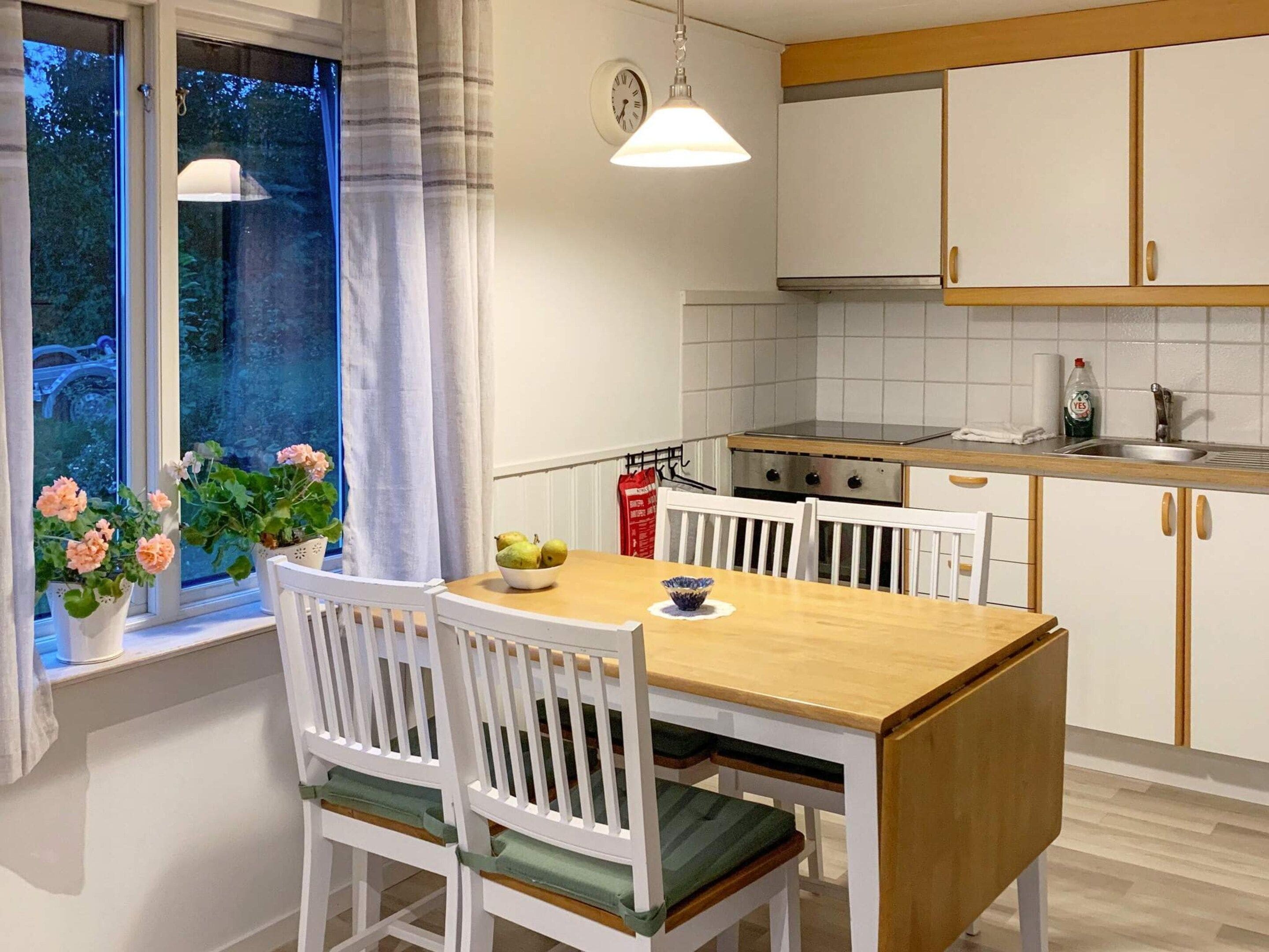 A kitchen with white cabinets, a wooden table, and chairs. There is a window with curtains and a view of greenery outside.