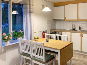 A kitchen with white cabinets, a wooden table, and chairs. There is a window with curtains and a view of greenery outside.