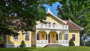 Ein gelbes Gebäude mit einem weißen Balkon und roten Türen, umgeben von grünem Gras und Bäumen.