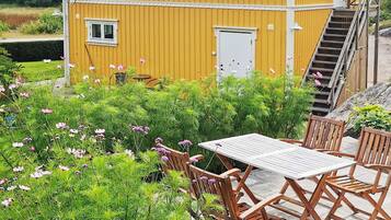 A yellow building with a staircase leading to a door, surrounded by lush greenery and a wooden table with chairs.