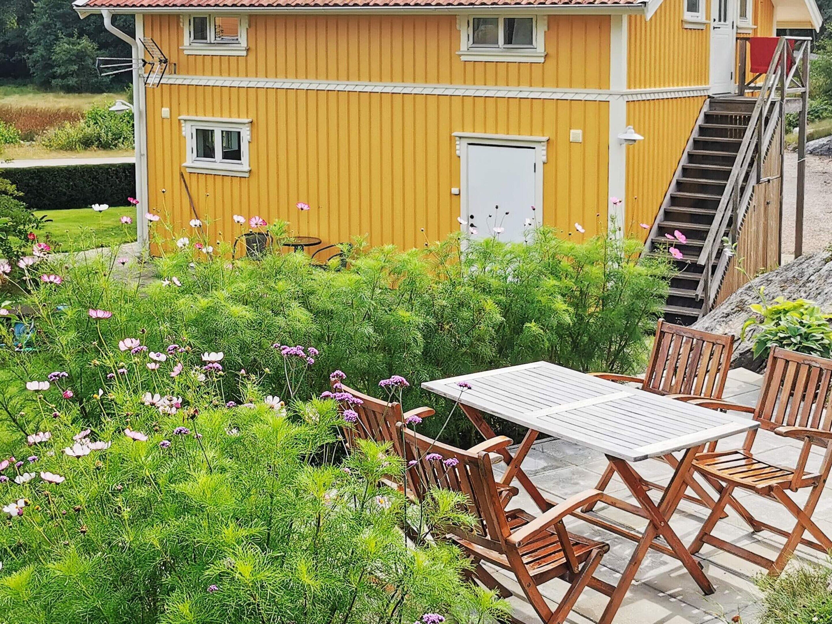 A yellow building with a staircase leading to a door, surrounded by lush greenery and a wooden table with chairs.