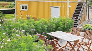 A yellow building with a staircase leading to a door, surrounded by lush greenery and a wooden table with chairs.