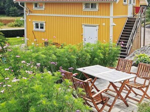 Um edifício amarelo com escadaria que leva a uma porta, cercado por vegetação exuberante e uma mesa de madeira com cadeiras.