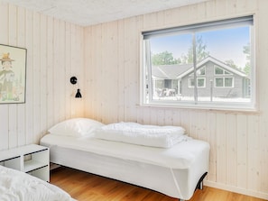 A bedroom with a bed, a bedside table, a wall-mounted lamp, and a window with a view of a house. - 5 Star Holiday Home in Vaeggerlose (Vaeggerlose)