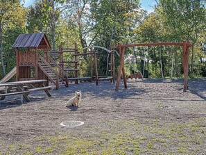 En lekplats med träkonstruktioner, inklusive en rutschkana och gungor, och en hund som sitter på marken.