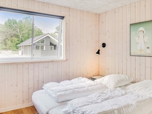 A bedroom with a large window, wooden walls, a bed with white bedding, a nightstand, and a wall-mounted lamp.