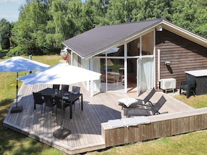 Una terraza de madera con mobiliario exterior, un espacio de patio cubierto y una sala con paredes de vidrio.