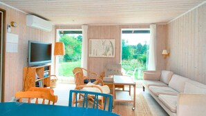A room with a television, wicker chairs, a sofa, and a large window offering a view of greenery.