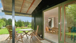 A wooden deck with a table and chairs, a glass door leading to an interior space with wicker furniture.