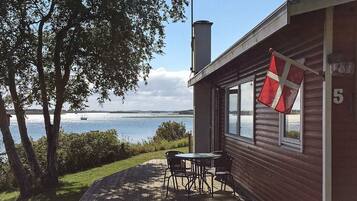 A wooden cabin with a flag, a deck with chairs, and a view of the water.