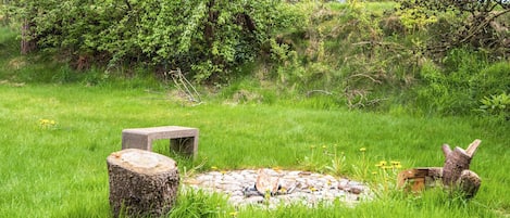 A natural outdoor setting with a stone bench, a tree stump, and a small wooden structure in a grassy area.
