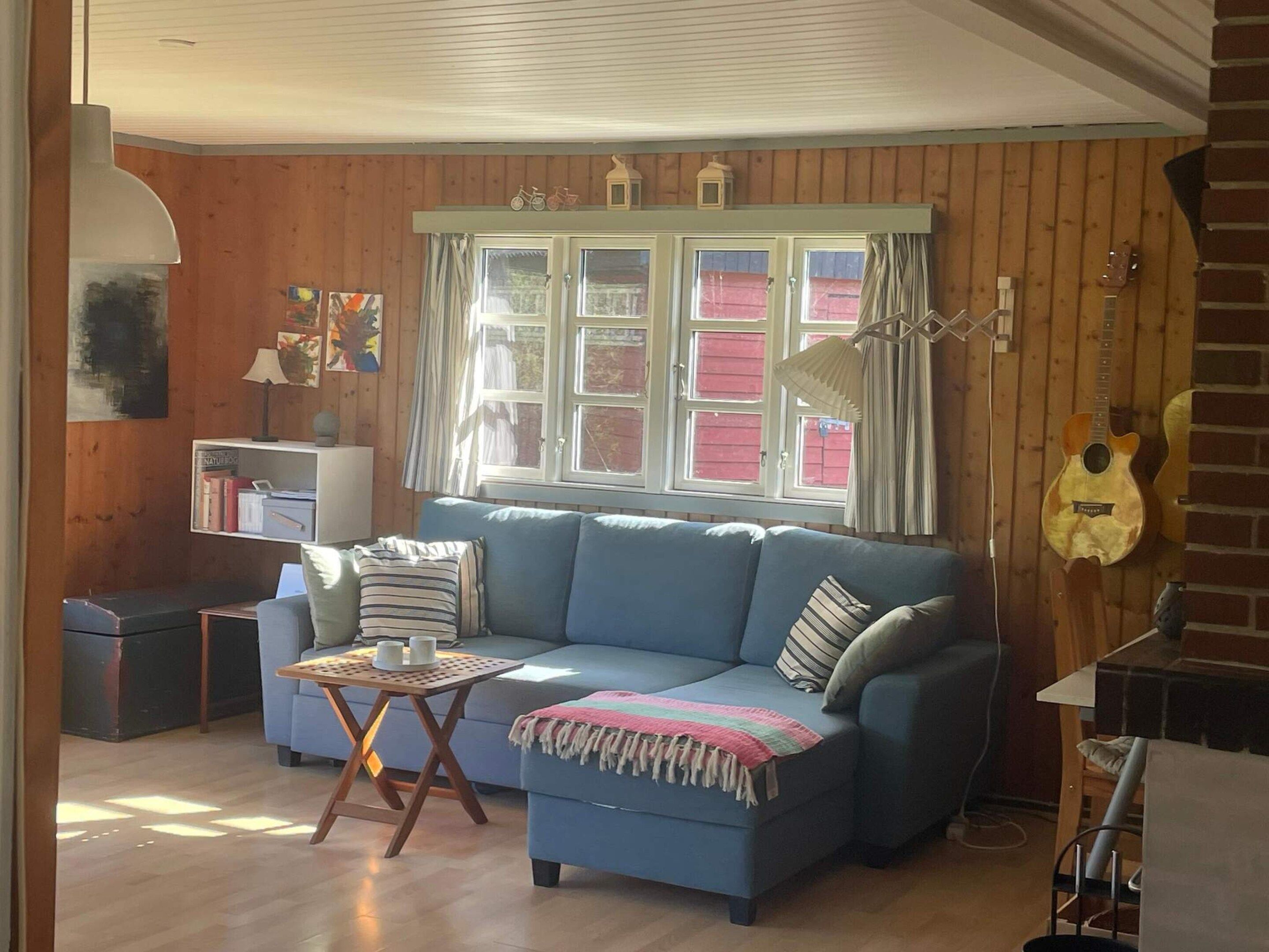 A cozy living room with a blue sofa, a small table with a lamp, a guitar, and a window with curtains.