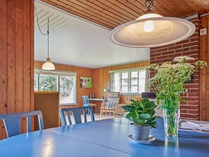 A dining area with a round table, chairs, a hanging light fixture, and a window with a view of greenery.