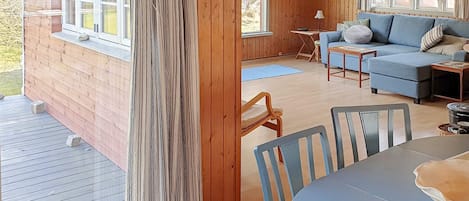 Interior de una cabaña de madera con comedor, sofá y ventana con vistas a un paisaje natural.