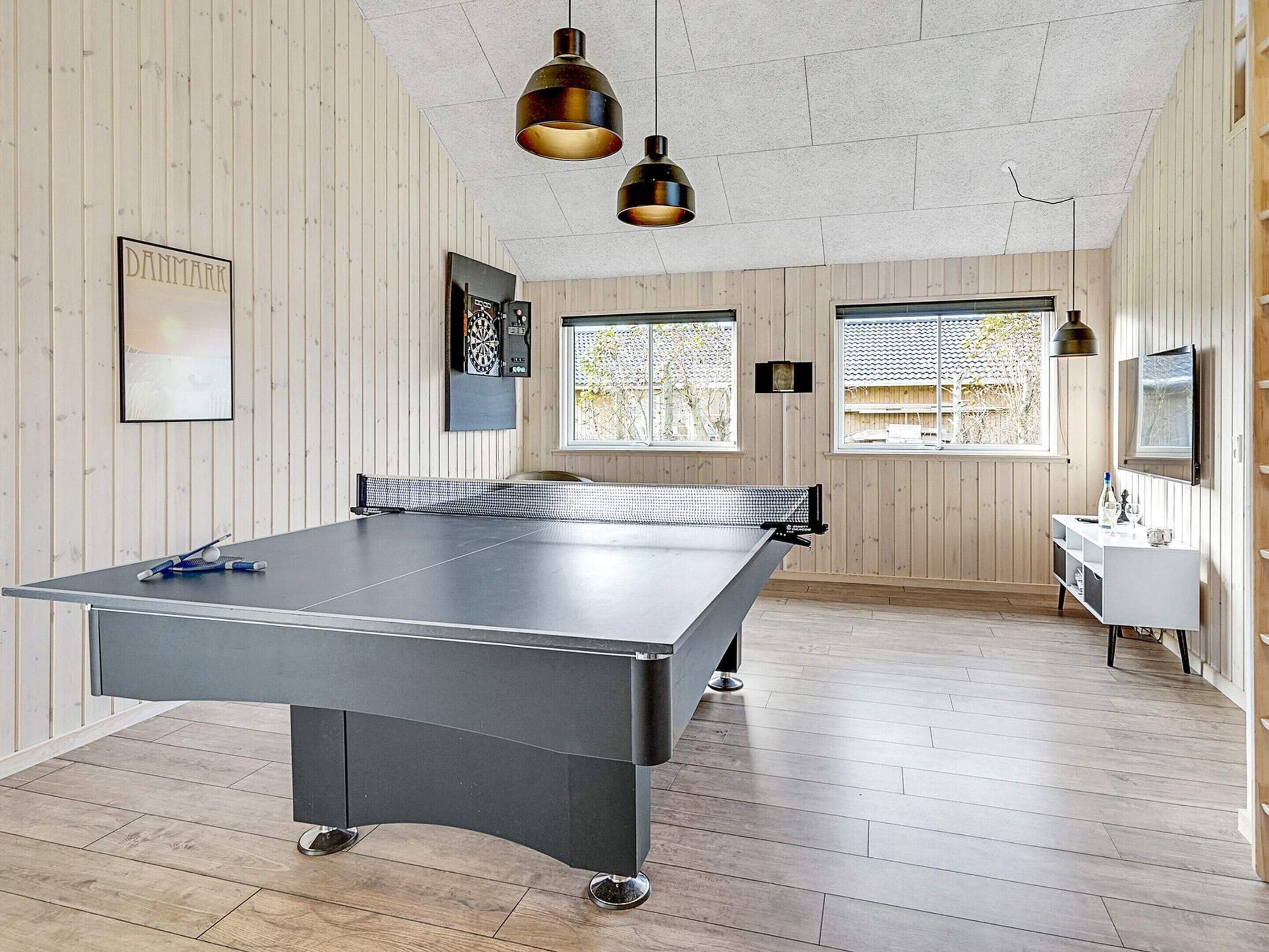 Mesa de pingue-pongue em um quarto com paredes de madeira, uma televisão e uma janela.