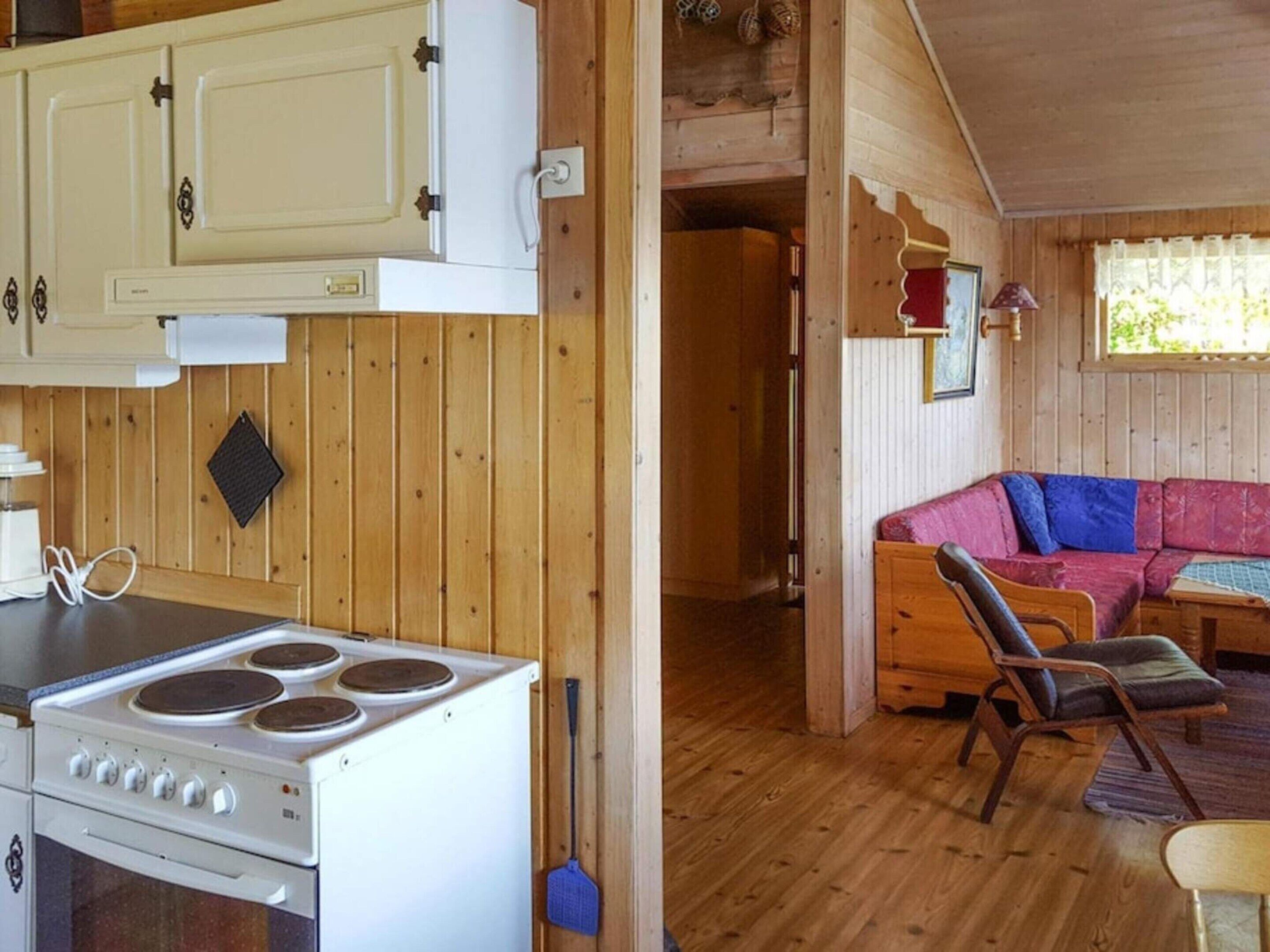 A wooden cabin interior with a kitchen area featuring a stove and cabinets, a living room with a sofa and chairs, and a window with curtains.