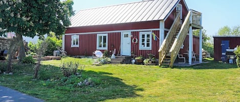 A red wooden house with a metal roof, a wooden staircase, and a small garden area.