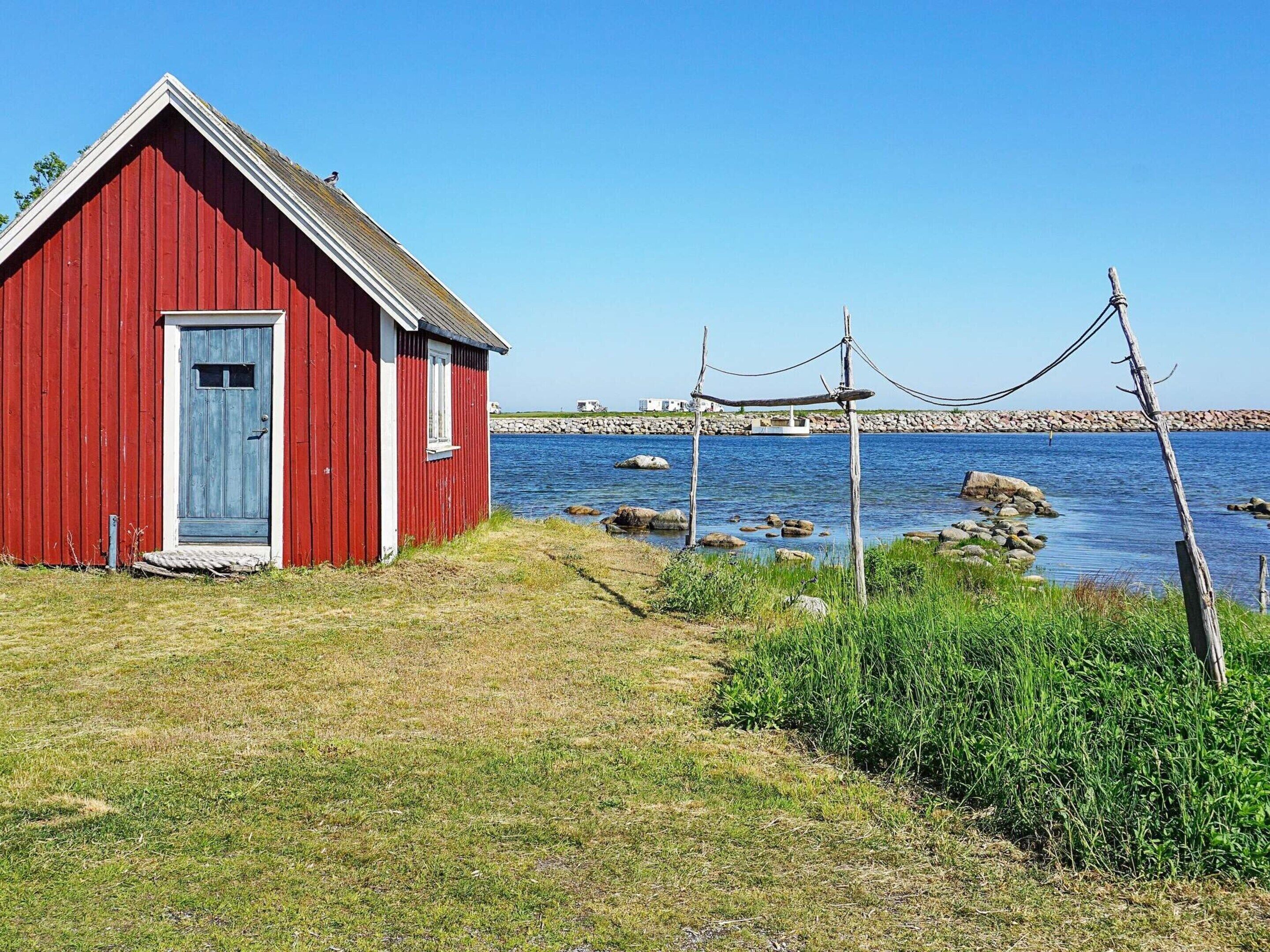 A red wooden building with a door and window, situated by a body of water with rocks and vegetation.