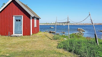 A red wooden building with a door and window, situated by a body of water with rocks and vegetation.