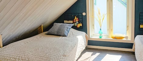 A bedroom with a sloped ceiling, a bed with a quilted cover, a window with a vase of dried plants, and a wall-mounted shelf with decorative items.