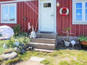 A red building with a white door, steps leading to the entrance, a life ring on the wall, and various outdoor items like a basket, umbrella, and potted plants.