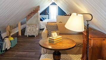 A cozy attic room with a sofa, a small table, and a lamp. There's a staircase leading to another room.