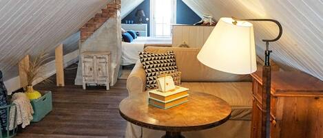 A cozy attic room with a sofa, a small table, and a lamp. There's a staircase leading to another room.