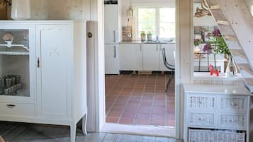 Una habitación con un armario blanco, una mesa de mimbre con cestas, y vista hacia una cocina con suelo de baldosas.