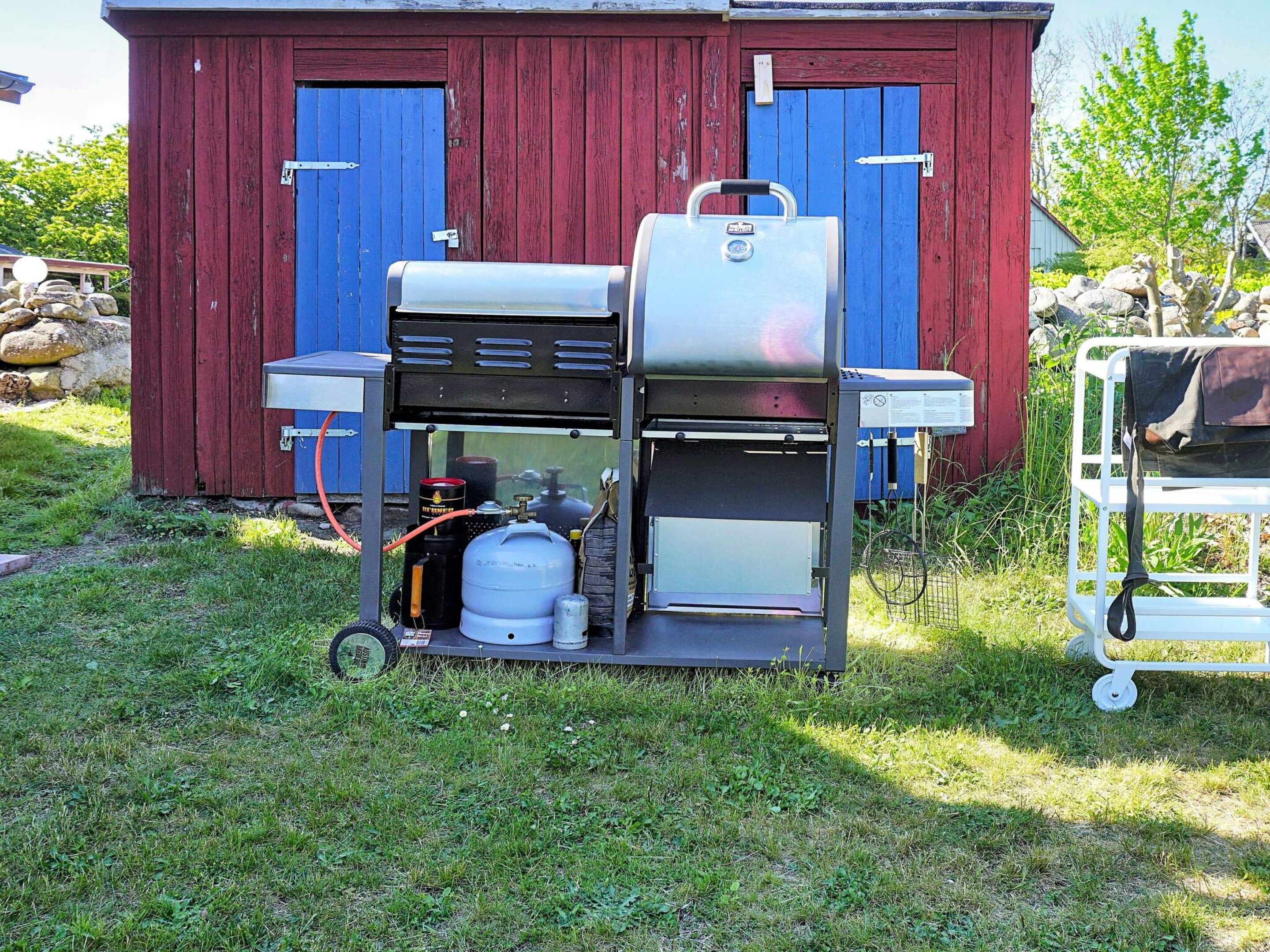 A portable gas grill with propane tanks and a cart, set up in front of a red and blue wooden building.