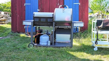 A portable gas grill with propane tanks and a cart, set up in front of a red and blue wooden building.