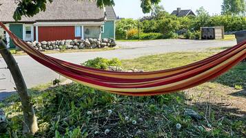 A hammock tied between two trees in a residential area with a house and a stone wall in the background.