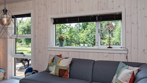 A modern living room with a grey sofa, colorful cushions, a wooden table, and a window with a view of greenery. - 5 Star Holiday Home in Vaeggerlose (Vaeggerlose)